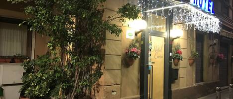 Entrada de hotel con un letrero que dice "Hotel Corso", plantas en macetas y una guirnalda decorativa de luces.