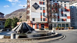 Un hotel con fachada moderna, un elemento acuático en primer plano y montañas al fondo.