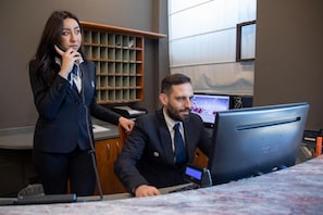 A person standing and talking on the phone, another person seated at a desk with a computer, and a shelf with multiple compartments in the background. - Vittoria (Sondrio)