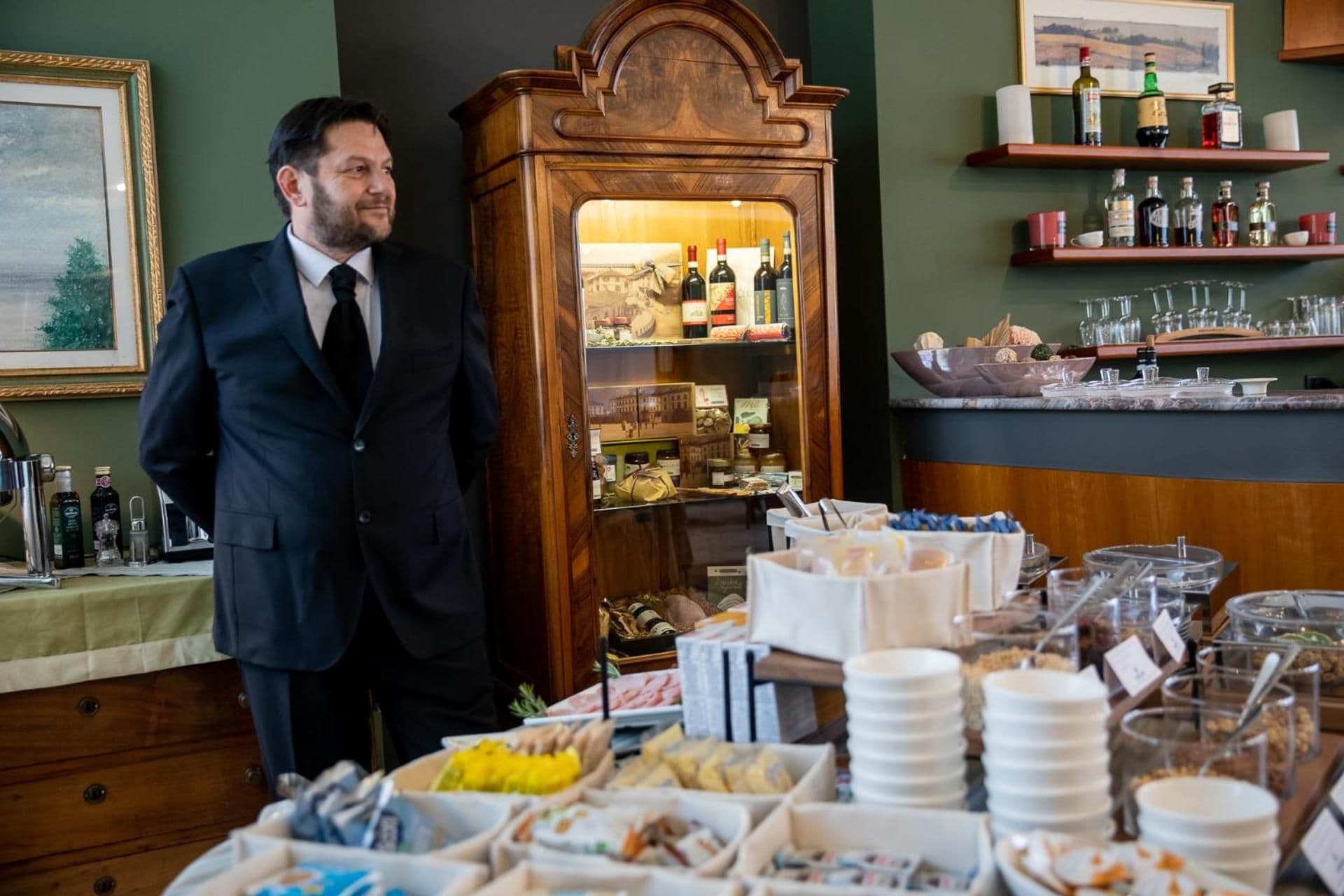 A man in a suit standing next to a wooden display cabinet filled with bottles, with a buffet table in the foreground.
