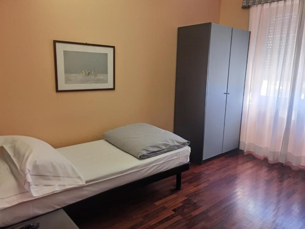 Una cama individual con colchón y almohadas, un armario, un cuadro en la pared y una ventana con cortinas.