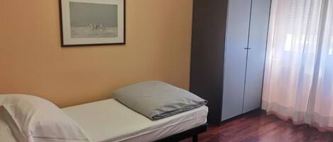 Una cama individual con colchón y almohadas, un armario, un cuadro en la pared y una ventana con cortinas.