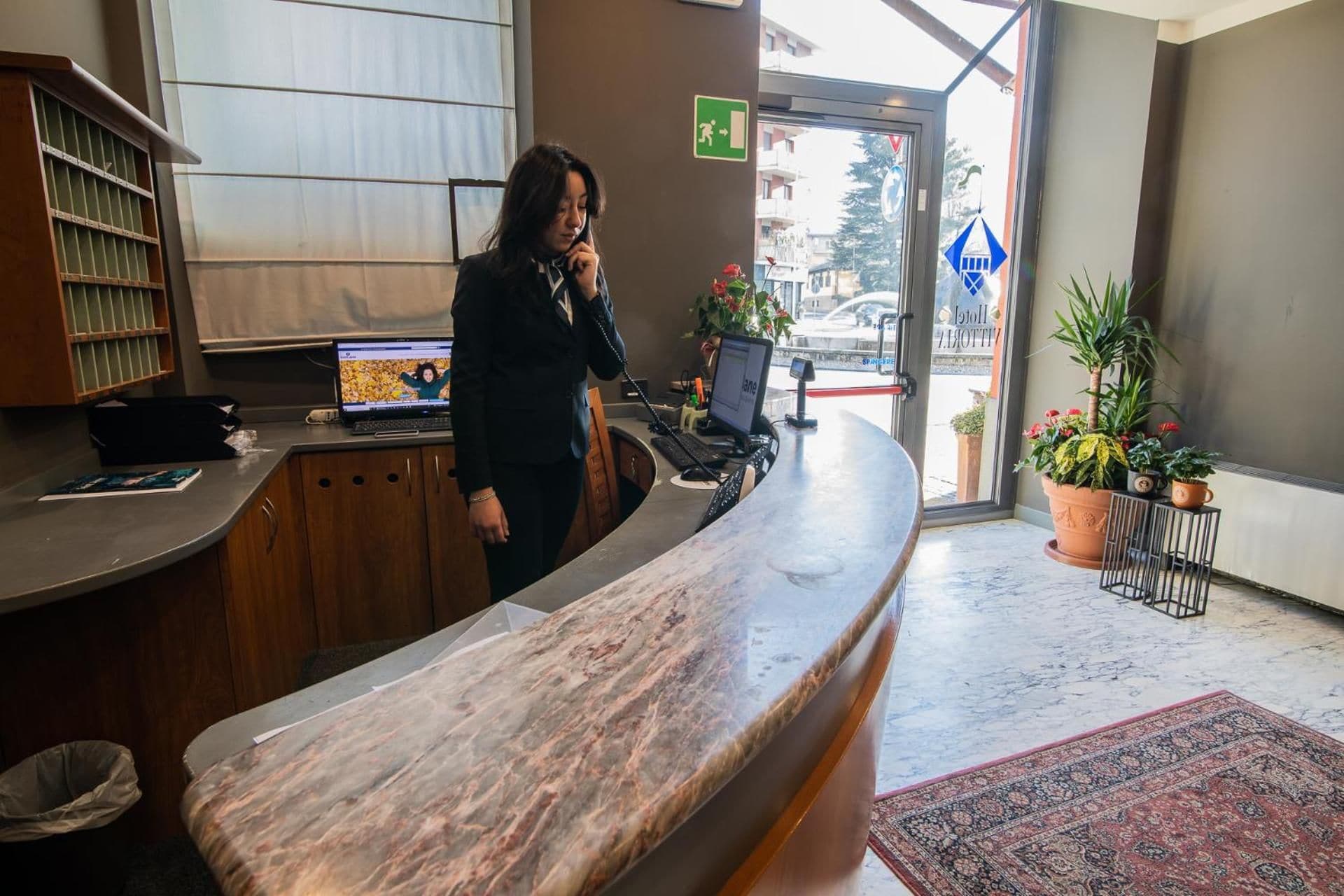 A reception desk with a computer, a telephone, and a plant. There is a window with a view of the street and a door with an emergency exit sign.