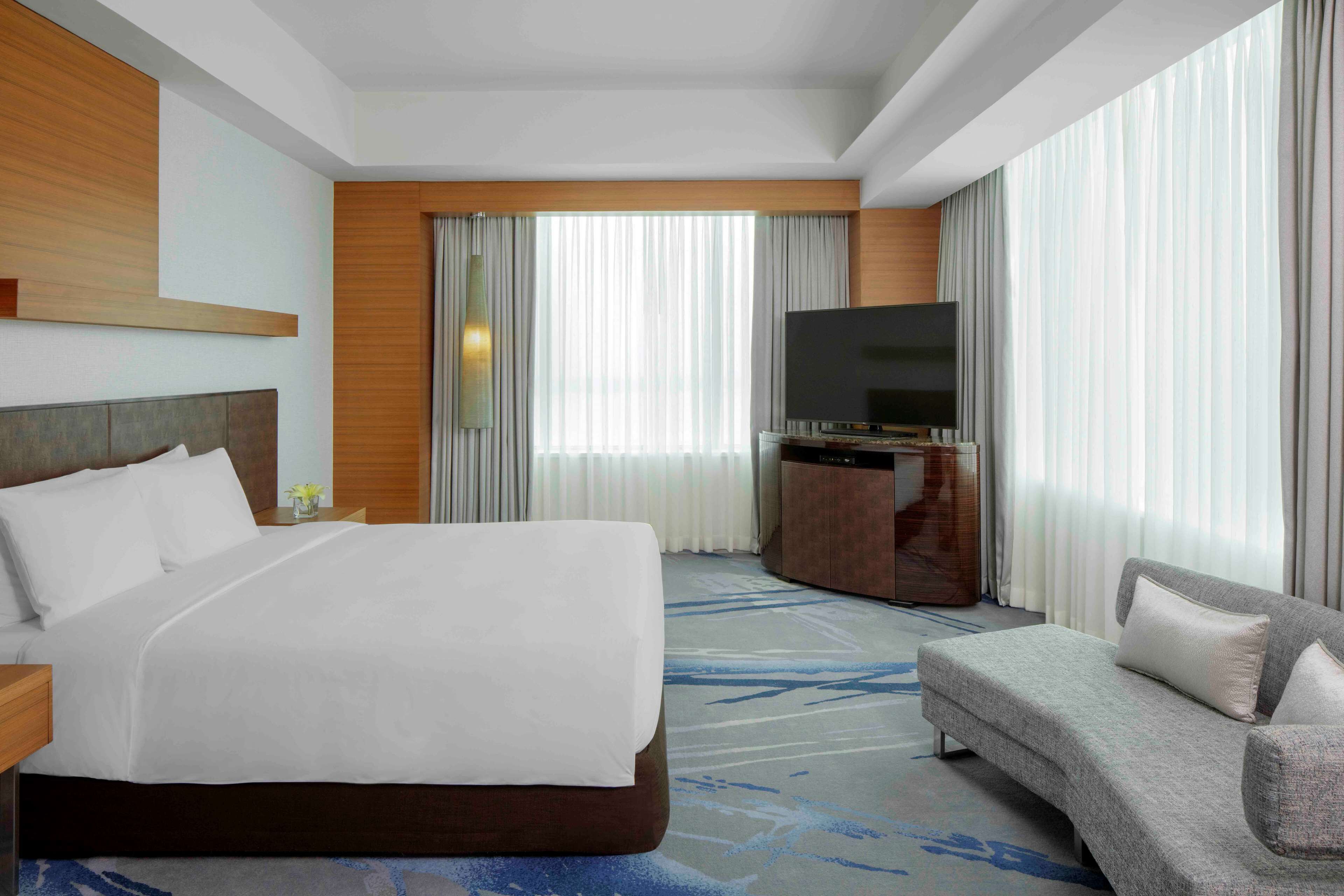 A hotel room with a large bed, a sofa, a flat-screen TV, and a large window with curtains.