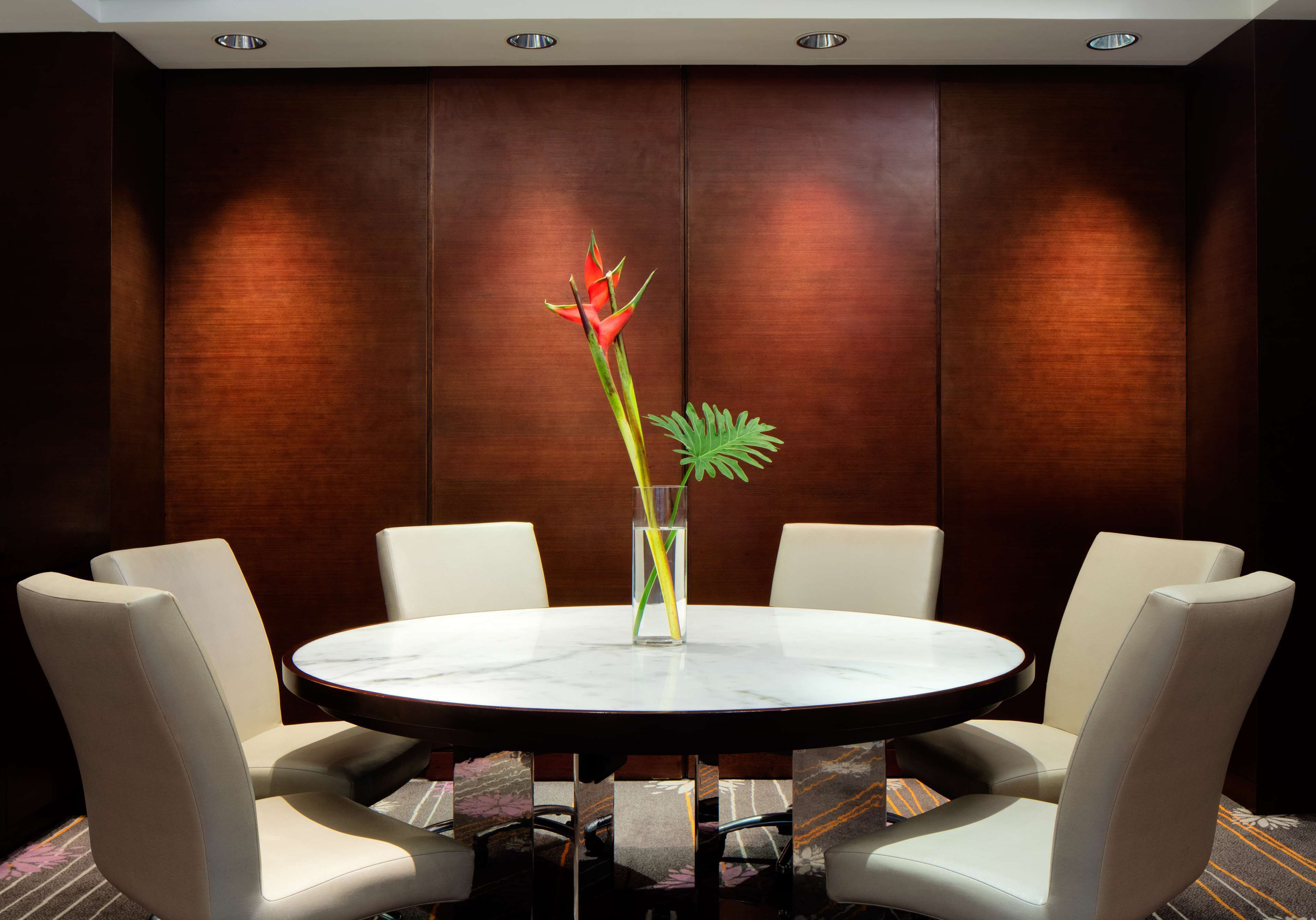 A round table with a marble top surrounded by six upholstered chairs. A tall vase with a single plant is placed on the table.