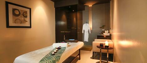 A modern spa room with a massage table, a small table with candles, and a robe hanging on a rack.