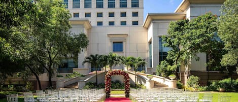An outdoor wedding venue with rows of white chairs, a red carpet, and a floral archway.
