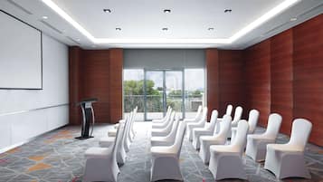 A conference room with rows of white chairs, a podium, and large windows offering a view of buildings and trees.
