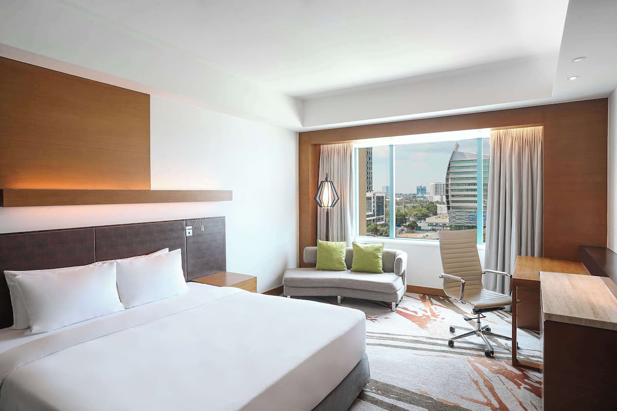 A modern hotel room with a large bed, a desk, a sofa, and a view of the city.