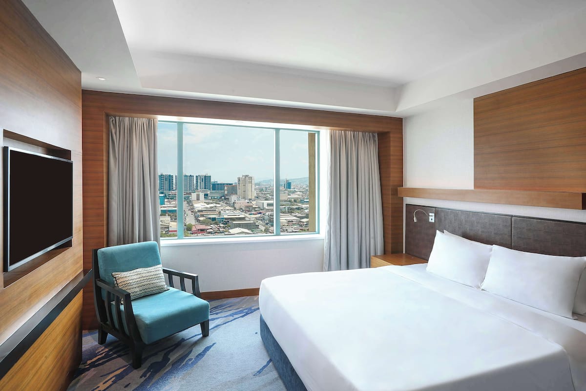 A hotel room with a large bed, a television, a chair, and a window with a city view.