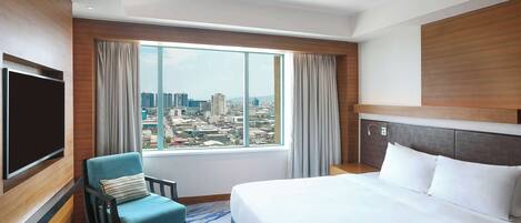 Ein Hotelzimmer mit einem großen Bett, einem Fernseher, einem Sessel und einem Fenster mit Blick auf die Stadt.