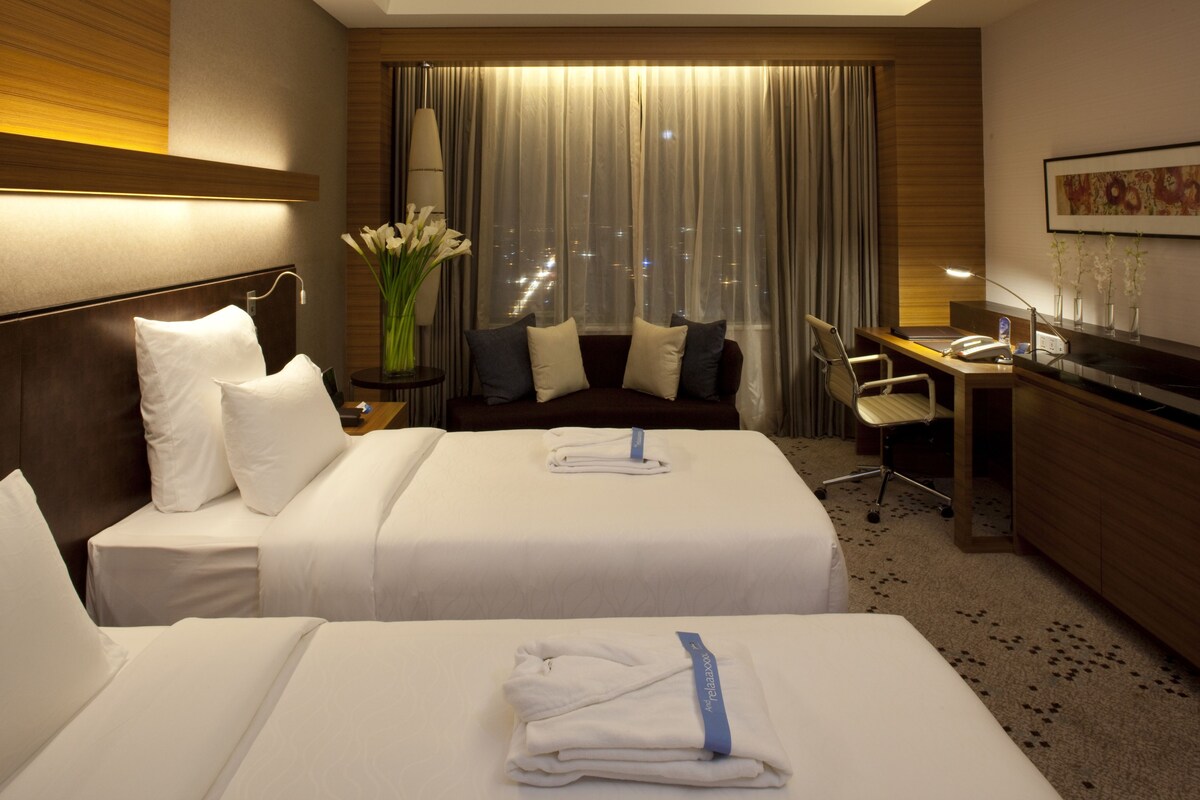 A hotel room with two beds, a desk, a chair, a lamp, a vase with flowers, and a city view through the window.
