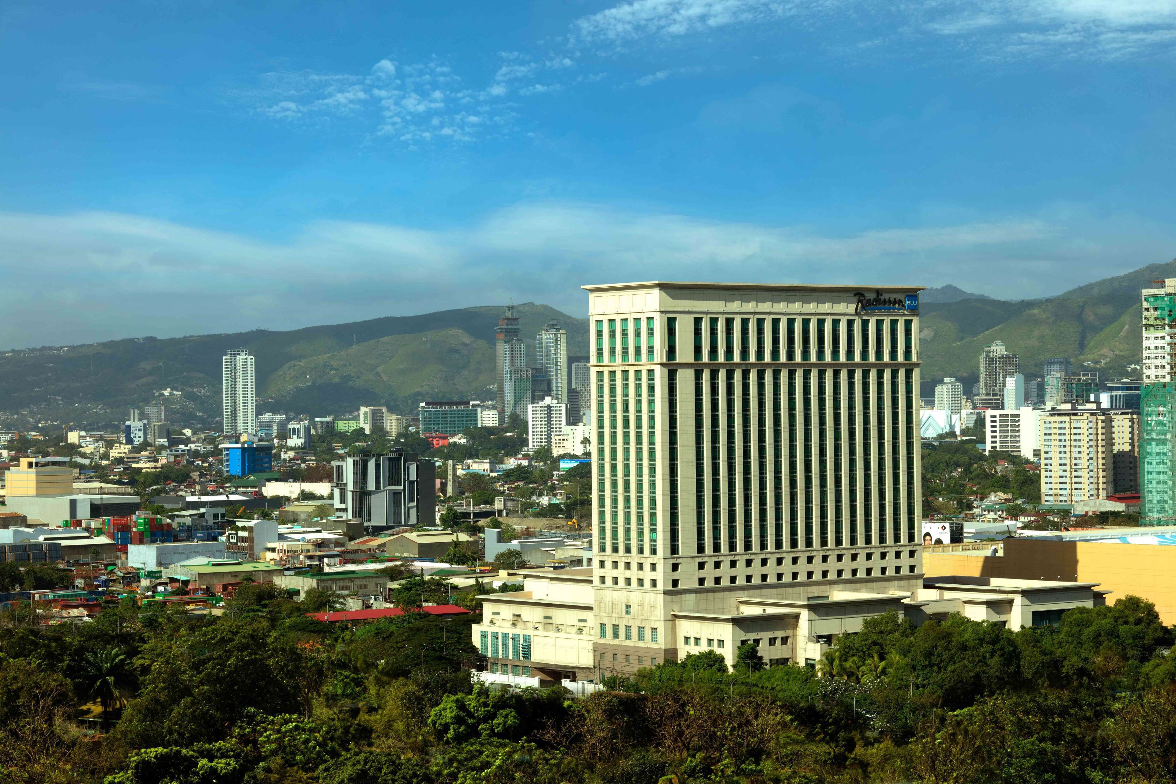 Un paisaje urbano con un edificio destacado de líneas verticales y techo plano, rodeado de otros edificios altos y áreas verdes.