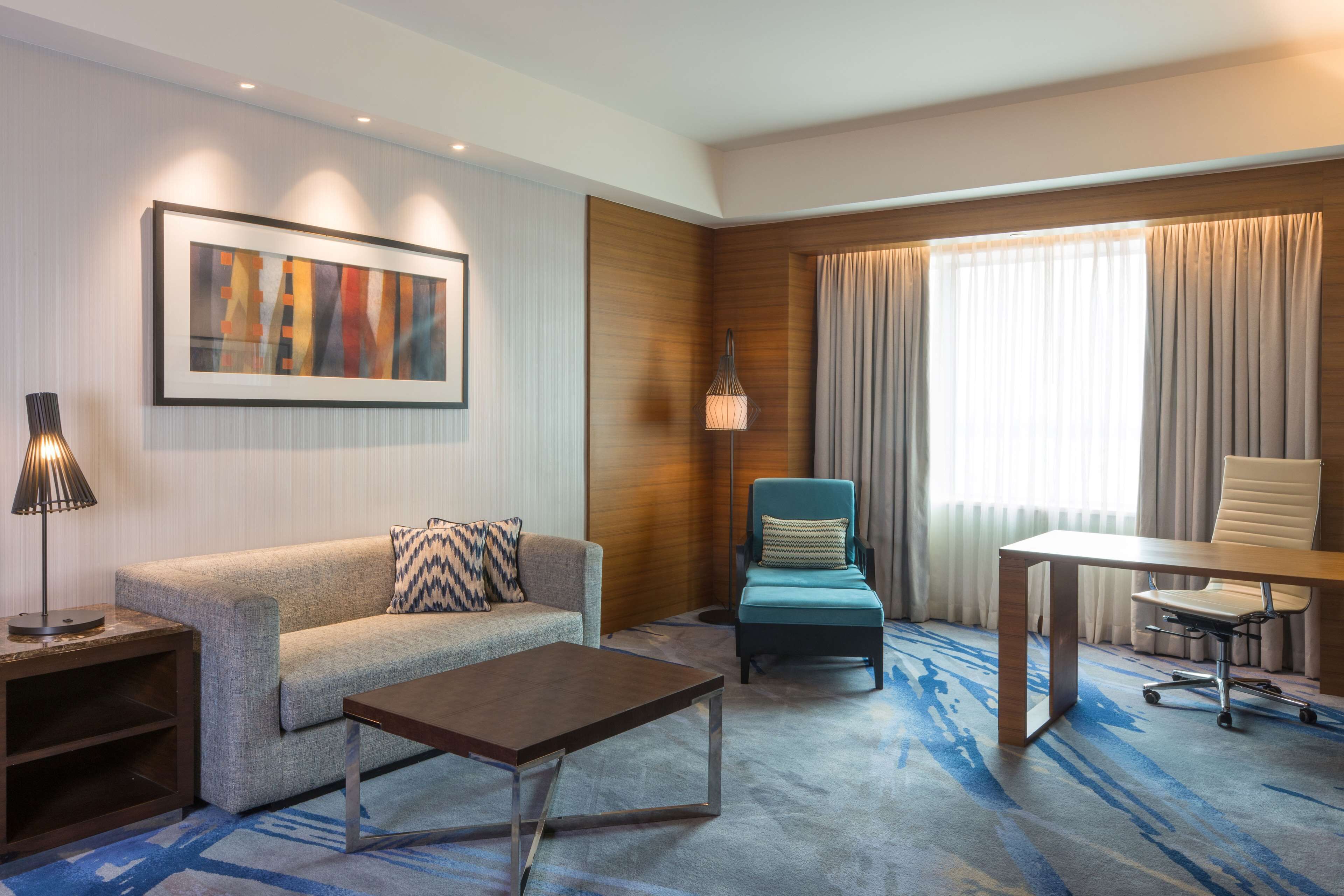 A modern hotel room with a sofa, a small table, a desk with a chair, a standing lamp, and a large window with curtains.