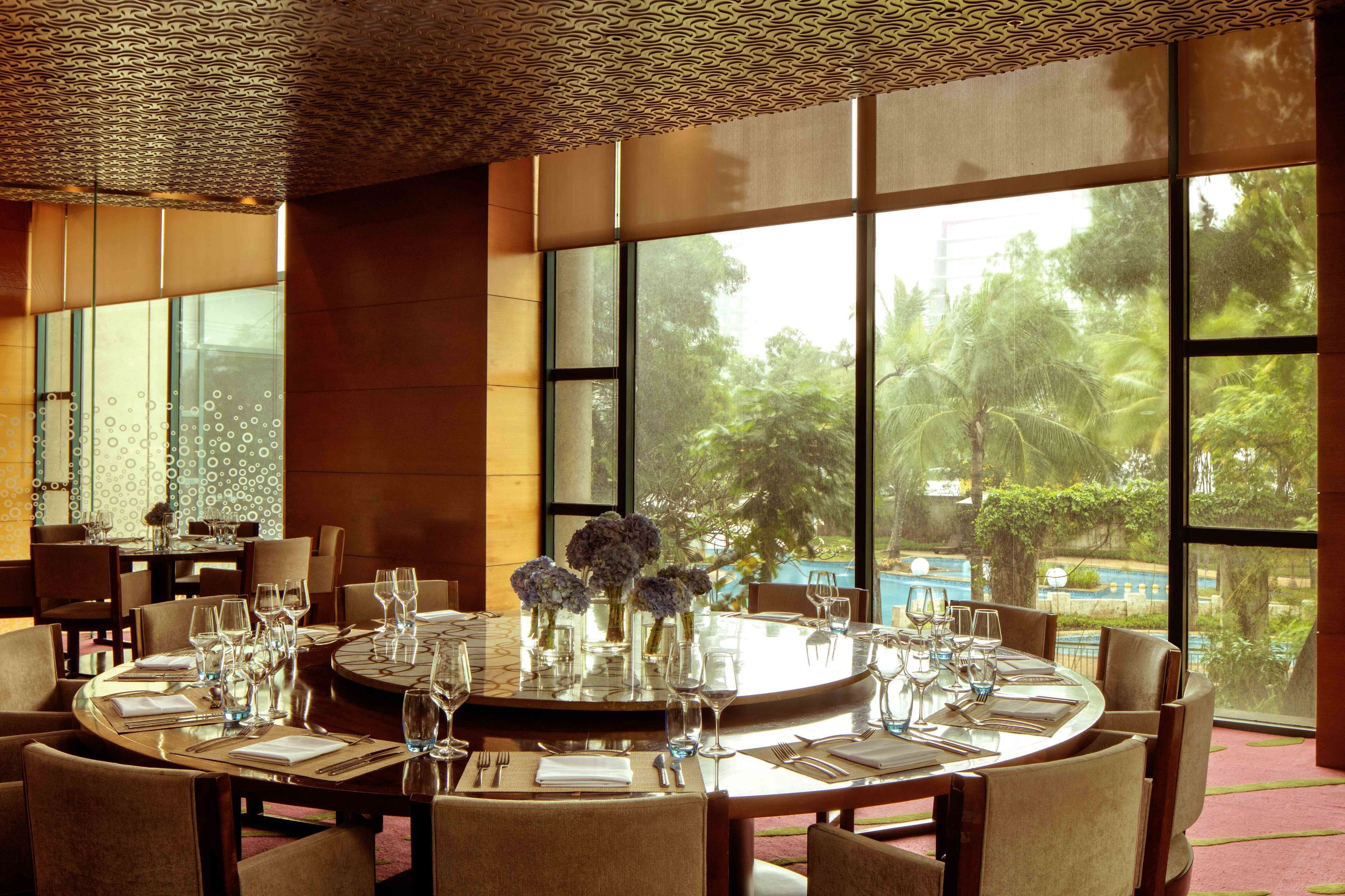 A dining area with round tables set for a meal, large windows offering a view of greenery and a pool, and a patterned ceiling.