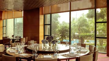 A dining area with round tables set for a meal, large windows offering a view of greenery and a pool, and a patterned ceiling.