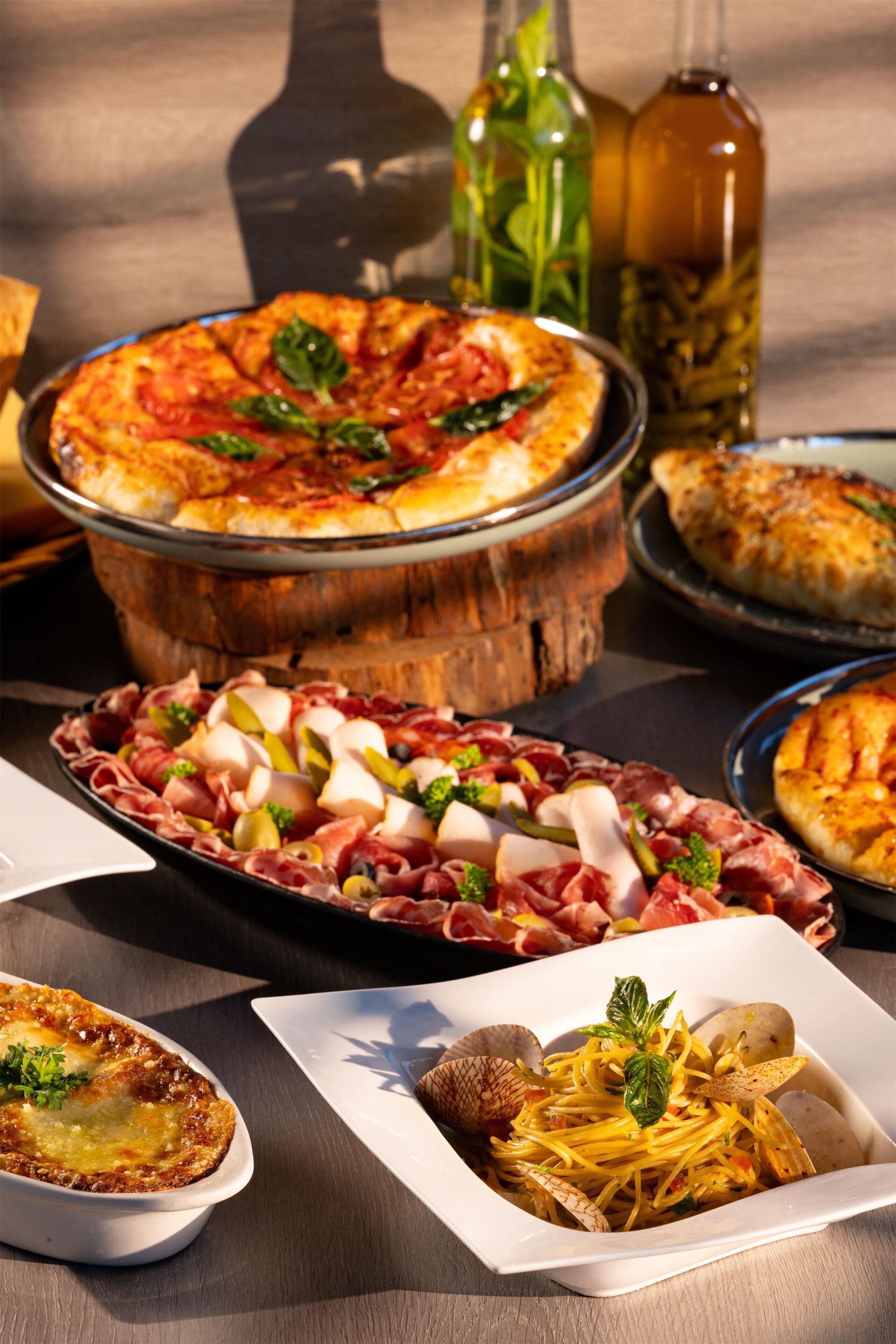 Forskellige retter, heriblandt pizza, pålæg og pasta, anrettet på et bord med flasker olivenolie i baggrunden.