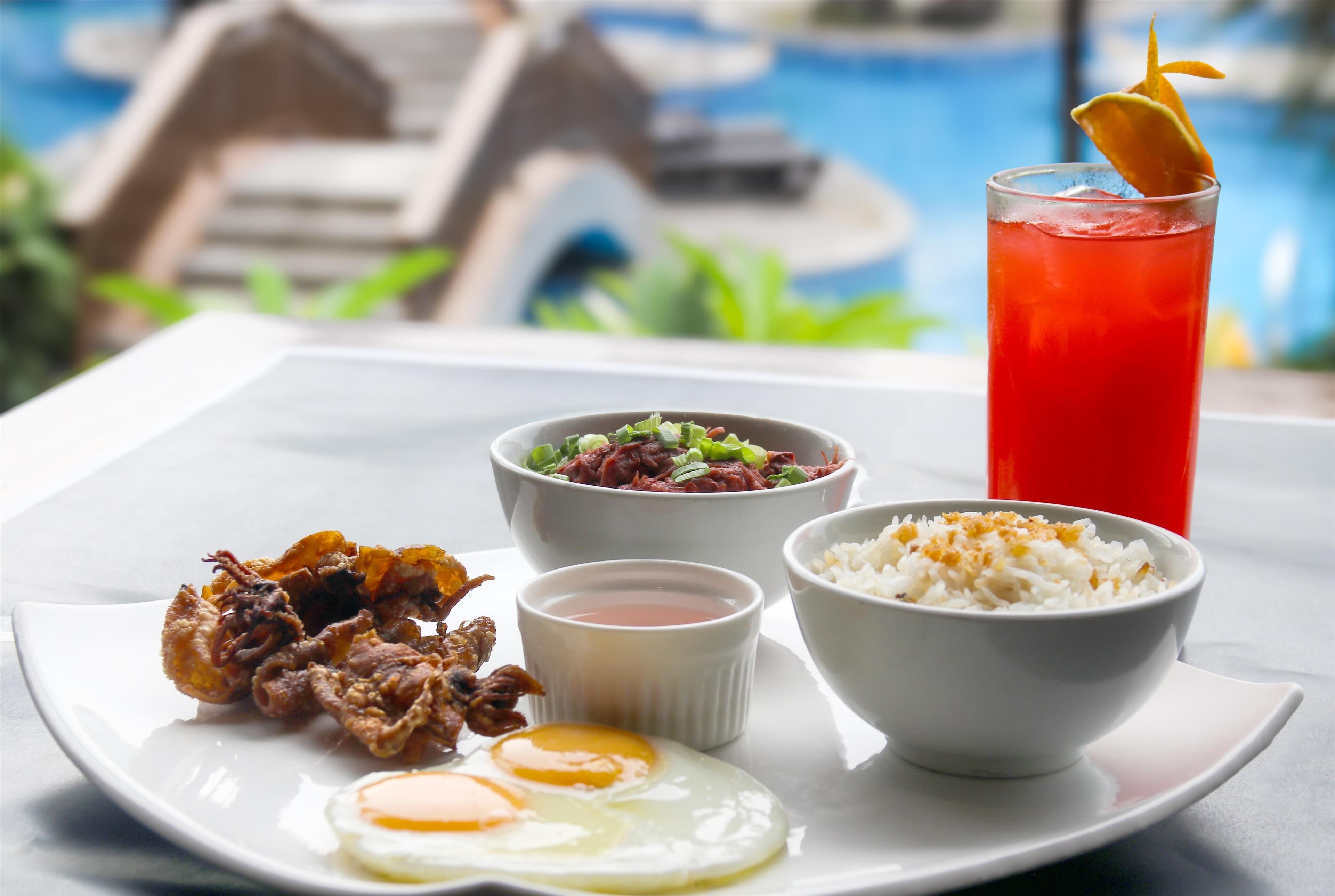Una mesa con un plato de pollo frito, arroz, dos huevos fritos, un tazón de ensalada y un vaso de bebida roja adornado con limón.