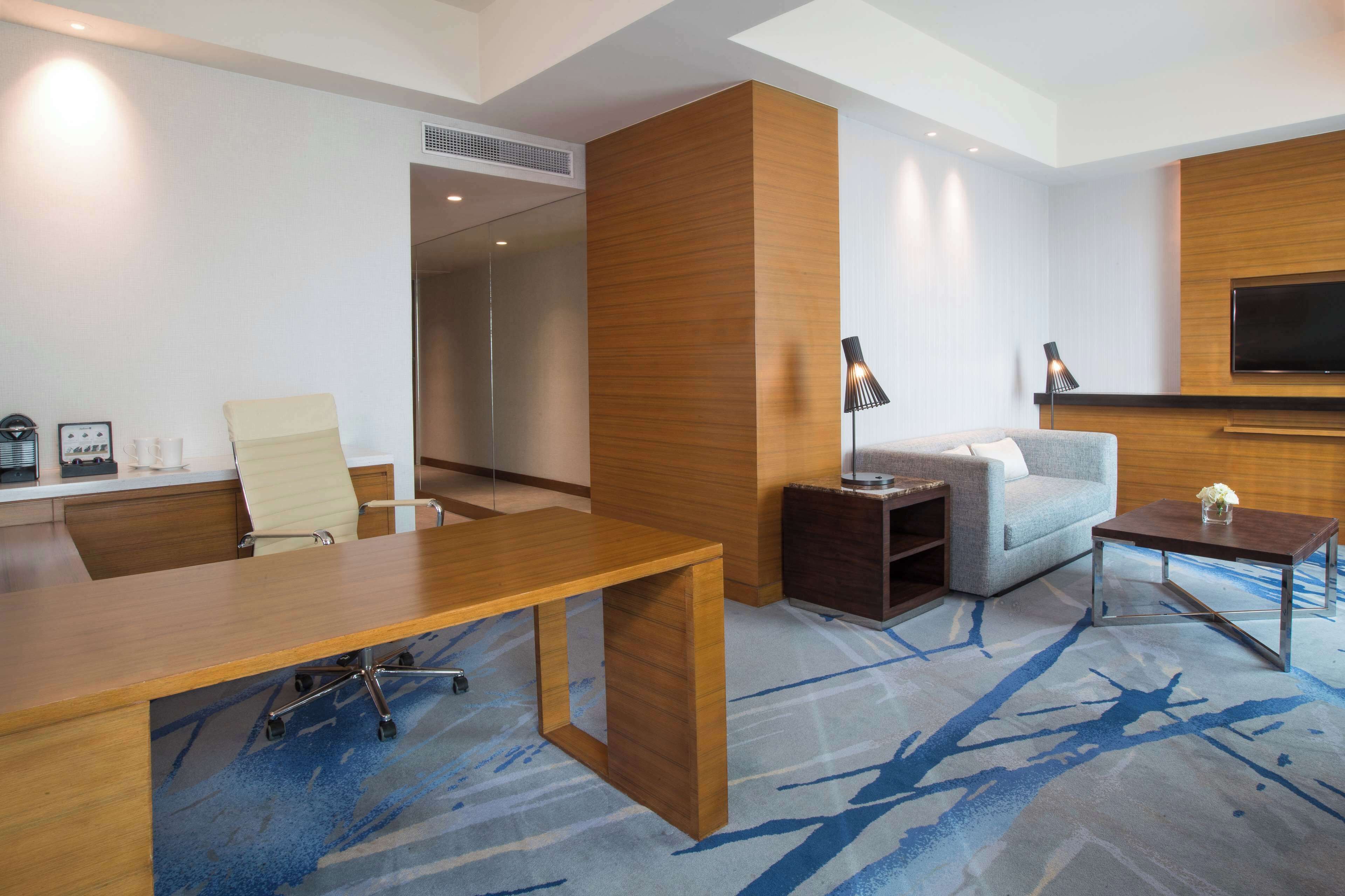 A modern hotel room with a wooden desk, a sofa, a TV mounted on the wall, and a patterned carpet.