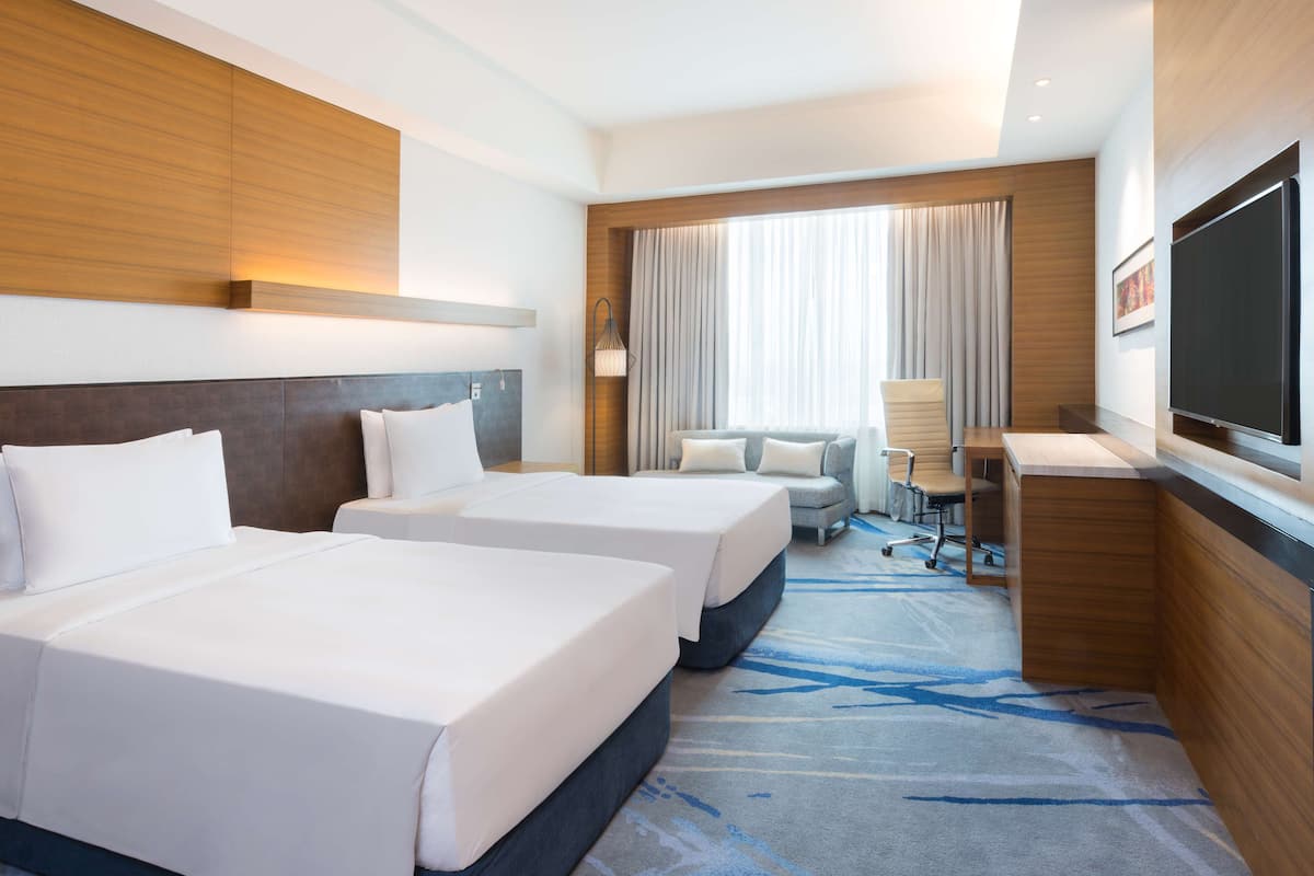 A hotel room with two beds, a desk, a chair, a TV, and a large window with curtains.