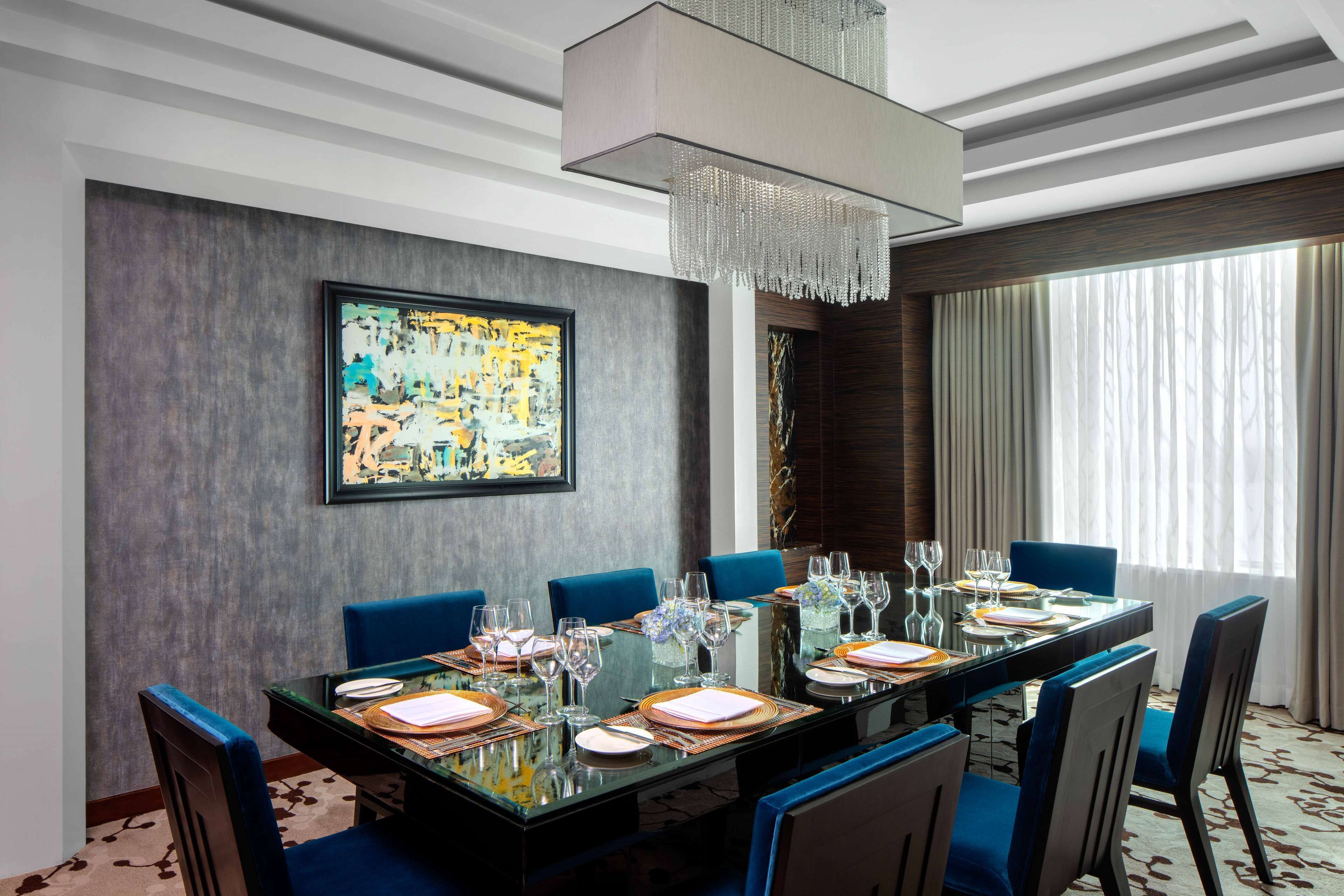 A modern dining room with a glass table set for six, blue chairs, a large abstract painting on the wall, and a chandelier.
