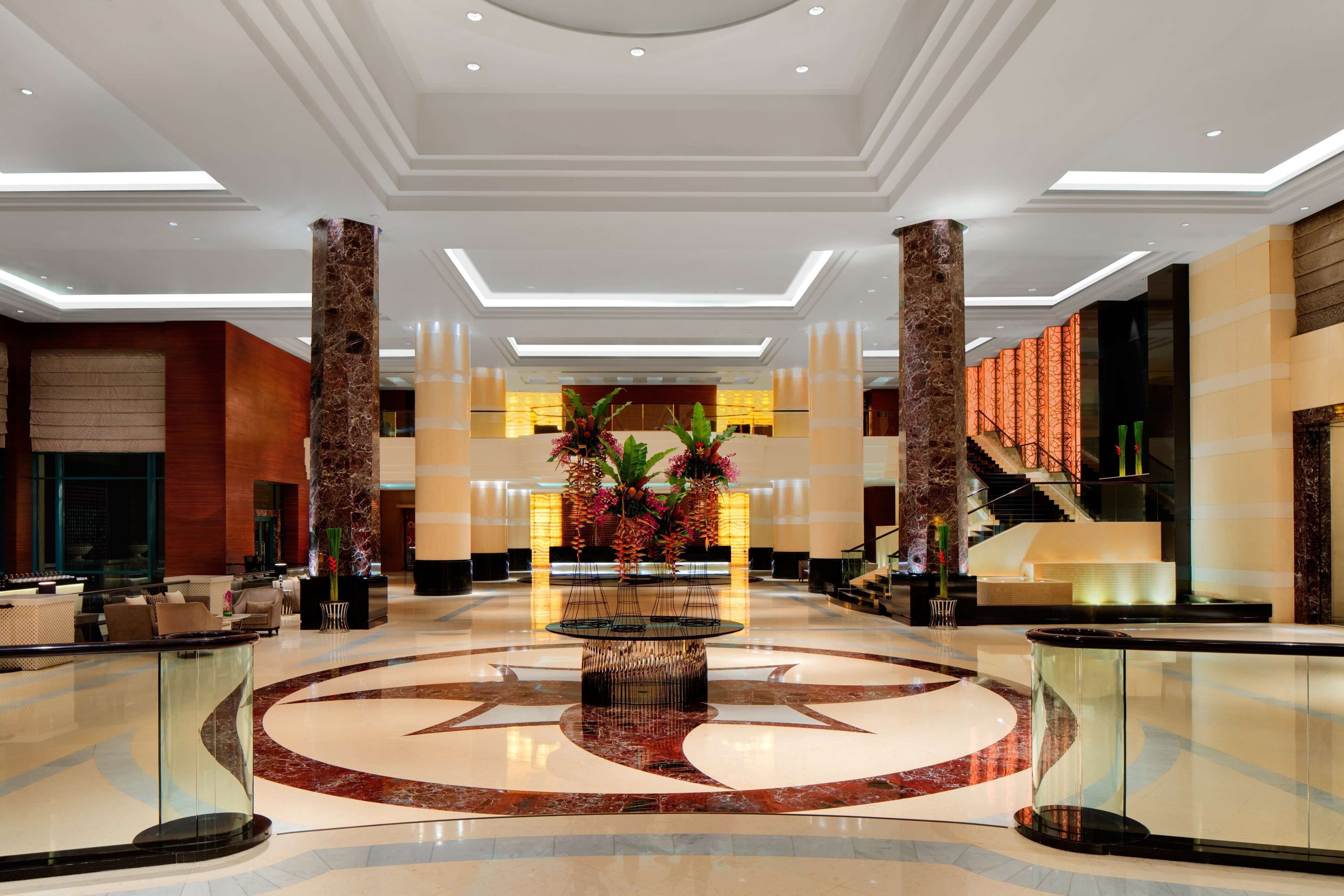 A spacious lobby with a central decorative fountain, a marble floor, and a staircase leading to upper levels.