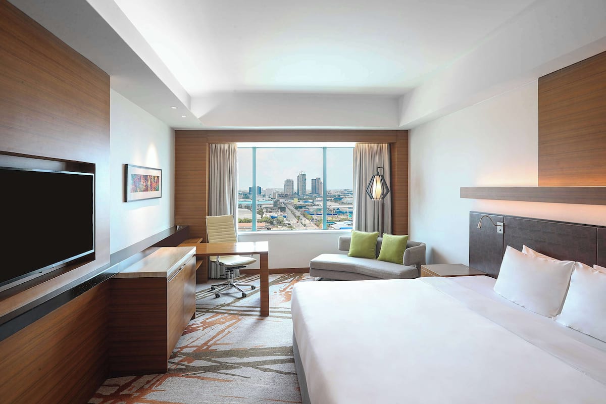 A hotel room with a large bed, a desk, a chair, a TV, and a window with a city view.