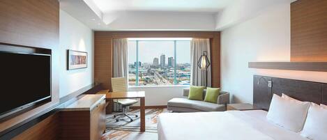 Ein Hotelzimmer mit einem großen Bett, einem Schreibtisch, einem Sessel, einem Fernseher und einem Fenster mit Blick auf die Stadt.