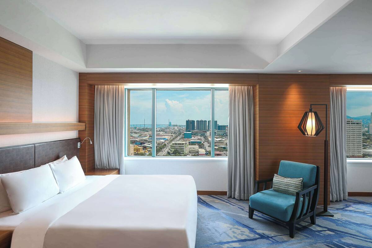 A hotel room with a large bed, a chair, a view of the city, and a lamp.