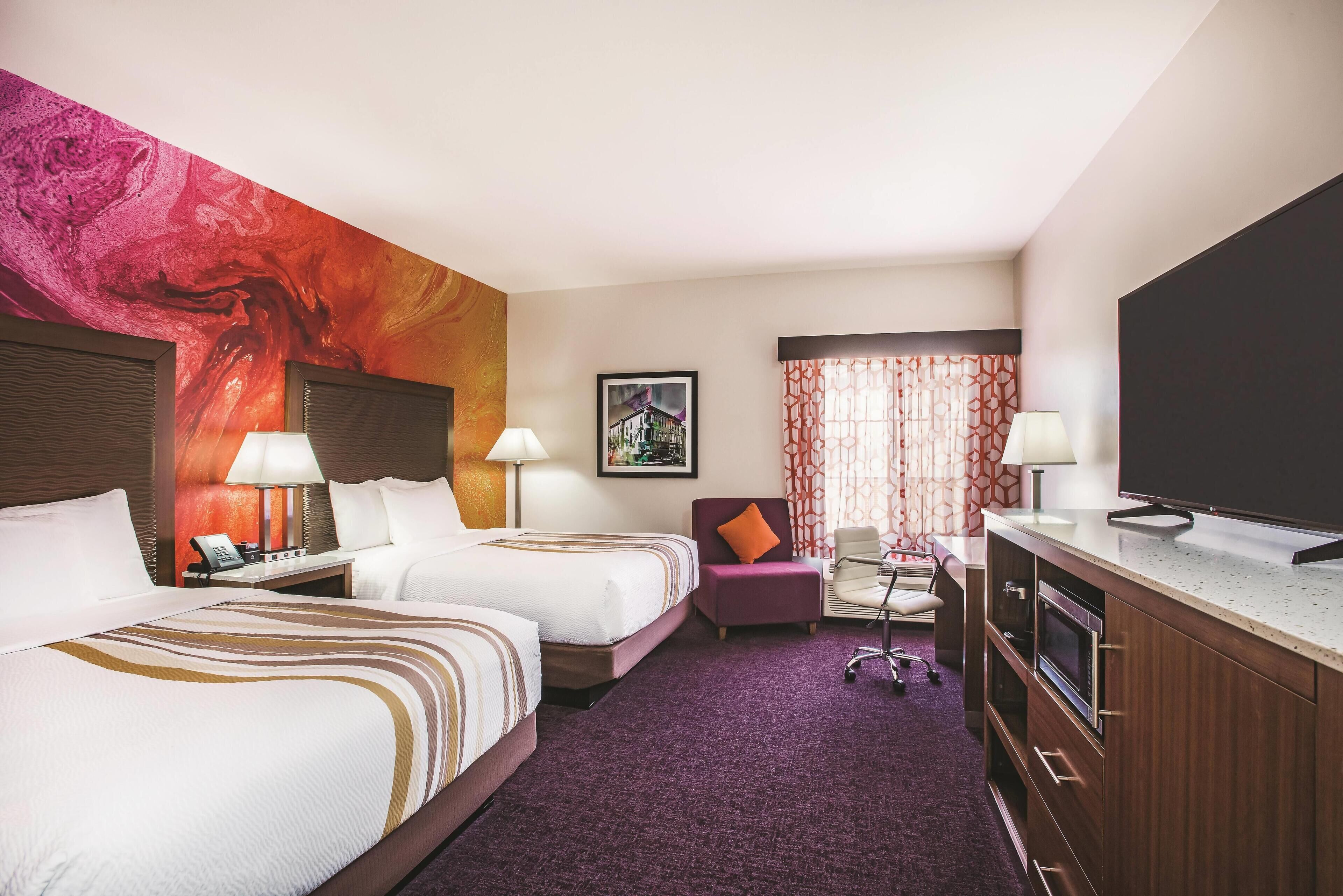 Habitación de hotel con dos camas, un escritorio, un televisor y un gran mural abstracto en la pared.