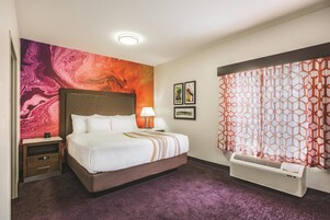 Habitación de hotel con una cama grande, una mesita de noche, una lámpara, una obra de arte en la pared, una ventana con cortinas estampadas y un equipo de aire acondicionado.