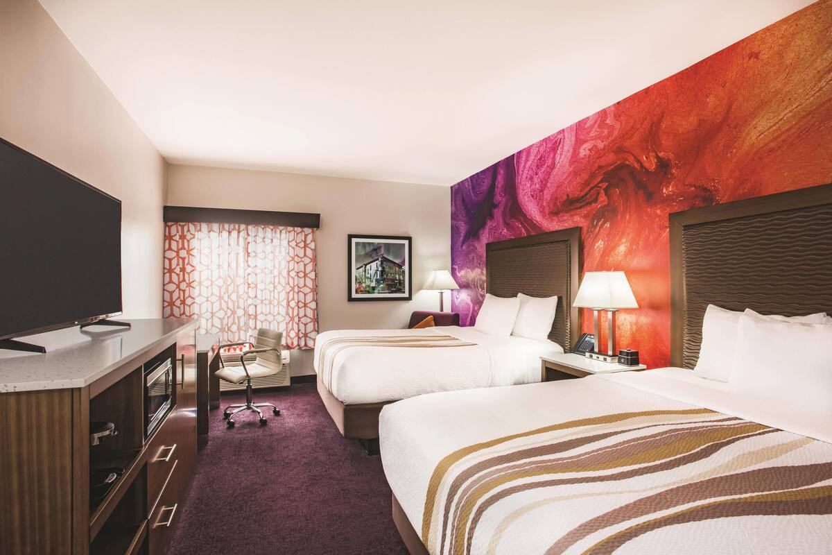 Une chambre d’hôtel avec deux lits, un bureau doté d’un téléviseur à écran plat, une chaise et un grand tableau abstrait accroché au mur.