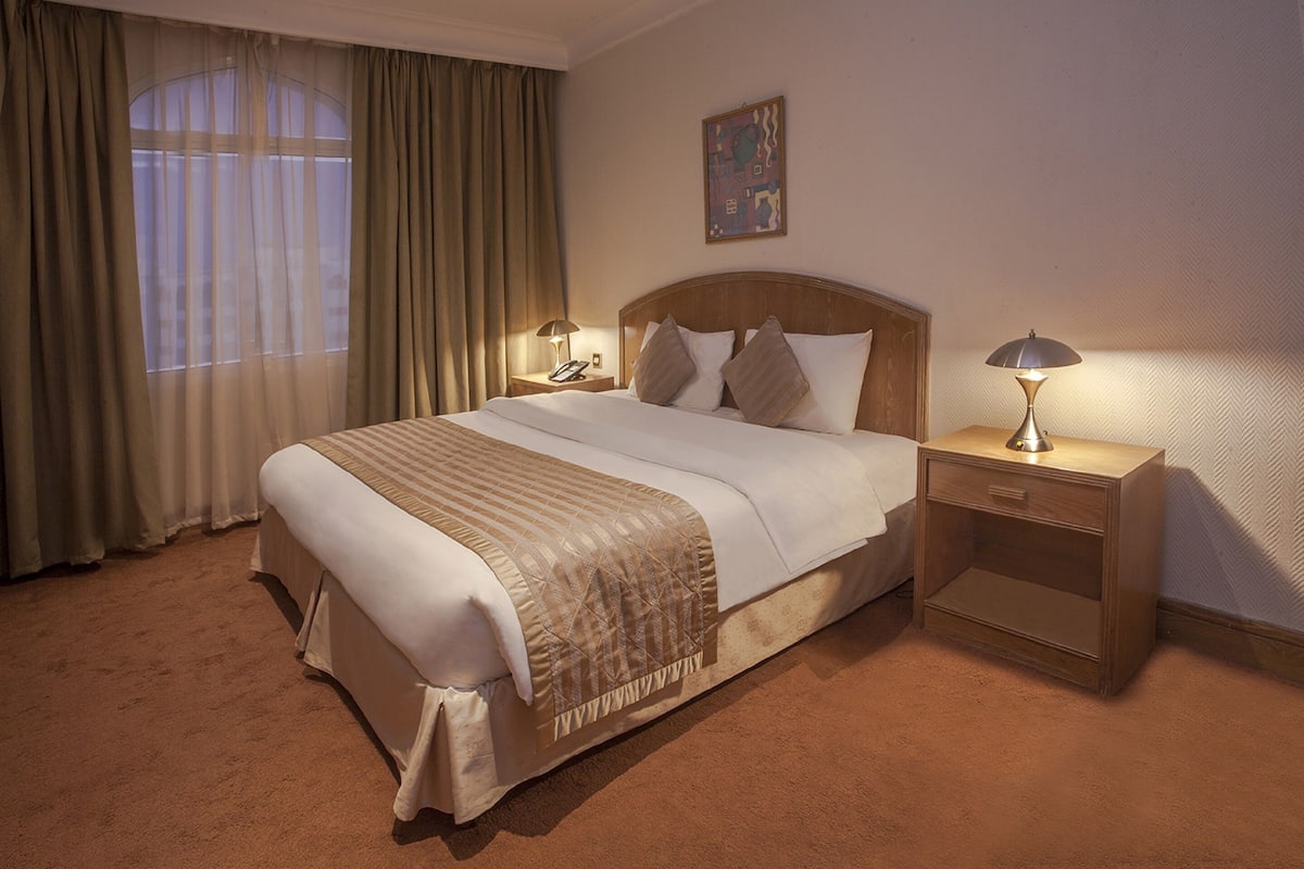 A hotel room with a large bed, two bedside tables with lamps, a window with curtains, and a painting on the wall.