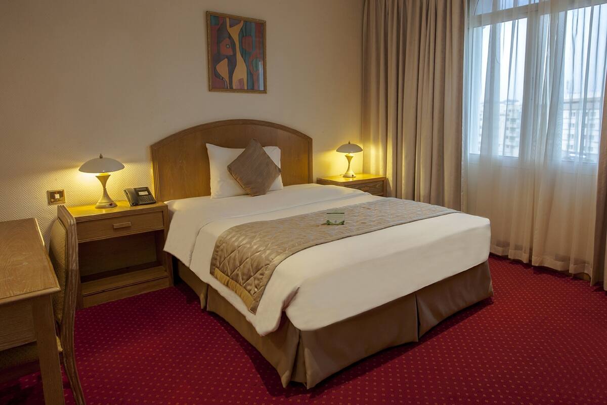 A hotel room with a large bed, two bedside tables with lamps, a wooden headboard, a red carpet, and a window with curtains.