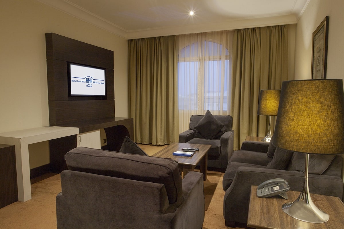 A hotel room with a flat-screen TV mounted on the wall, a sofa set, a wooden coffee table, a lamp, and a telephone on a side table.