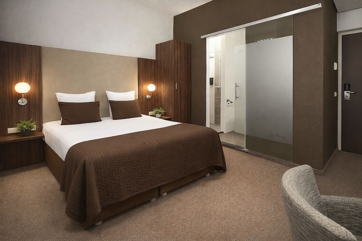 Quarto de hotel moderno com uma cama grande, duas mesas de cabeceira com luminárias, um guarda-roupa e zona de duche.
