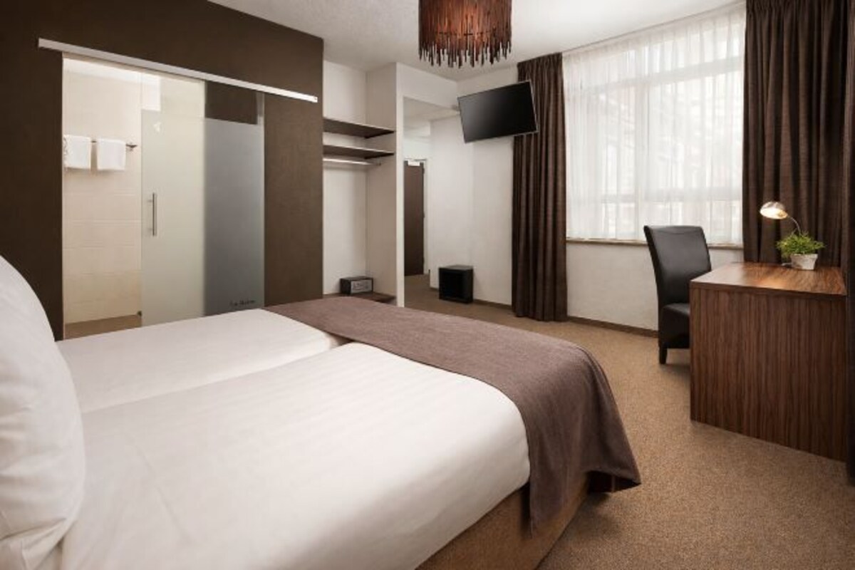 A hotel room with a large bed, a desk, a chair, a TV, and a window with curtains.