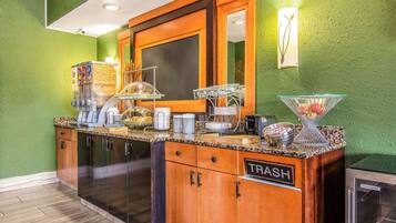 Zona de buffet con un contenedor de basura etiquetado "TRASH", una máquina de café y un bol de fruta.
