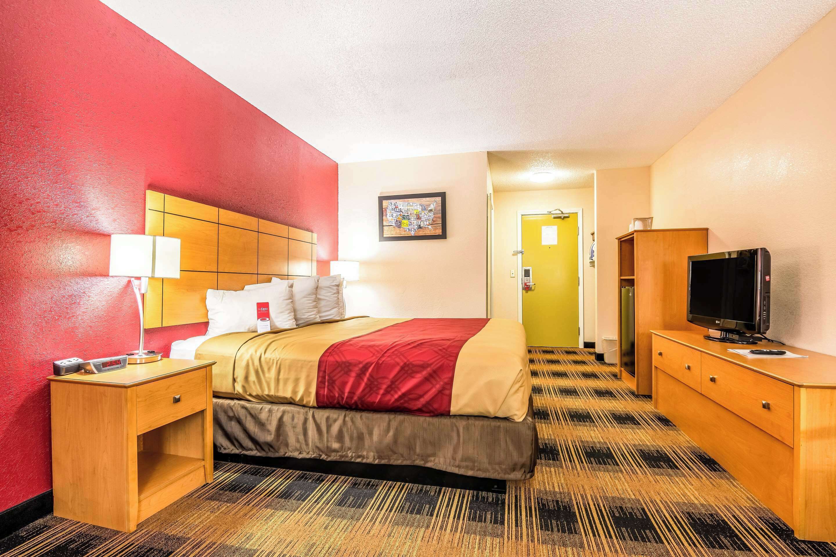 A hotel room with a bed, bedside table, lamp, TV, and a door leading to another room.