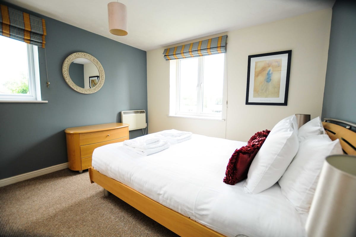 A neatly made bed with white linens, a wooden nightstand, a round mirror, a radiator, and a window with curtains.