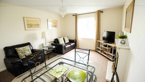 A living room with a black leather sofa, a glass coffee table, a TV on a wooden stand, and a chandelier. - Your Stay Bristol Orchard Gate (Bristol)