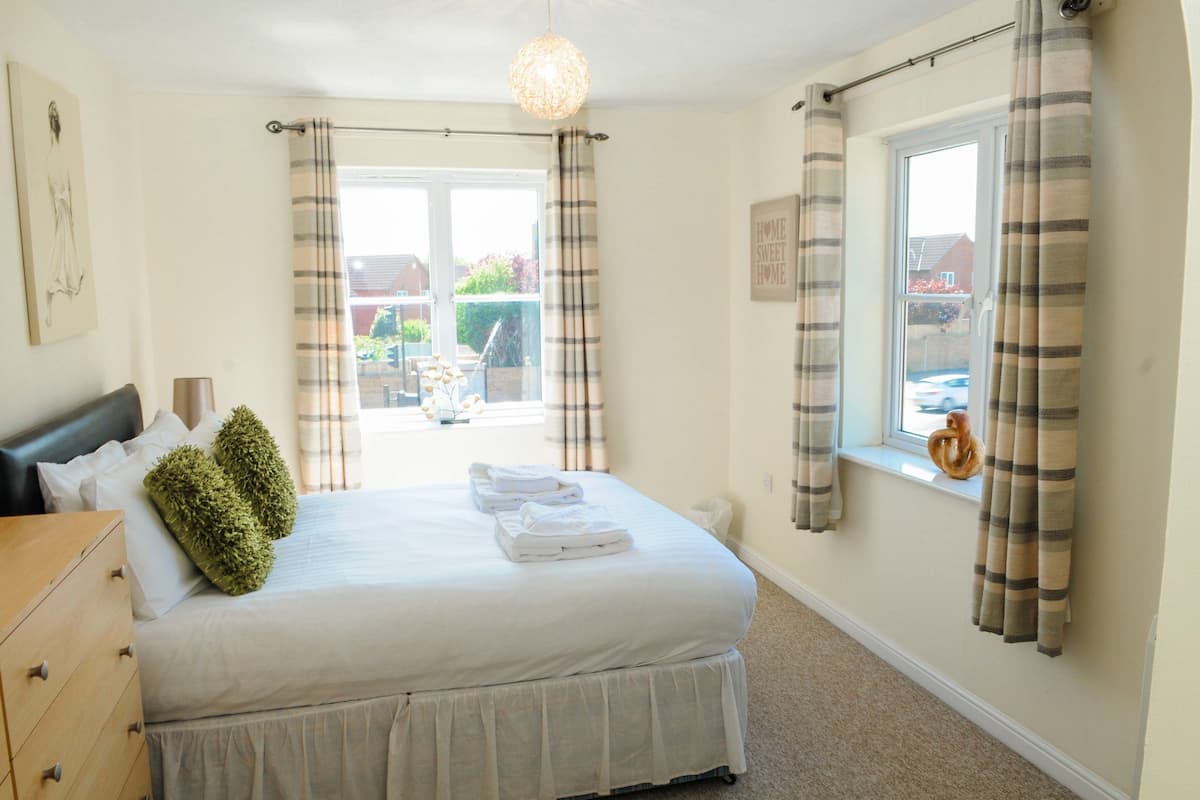 A neatly made bed with pillows and folded towels, a wooden dresser, a window with curtains, and a view of the outside.