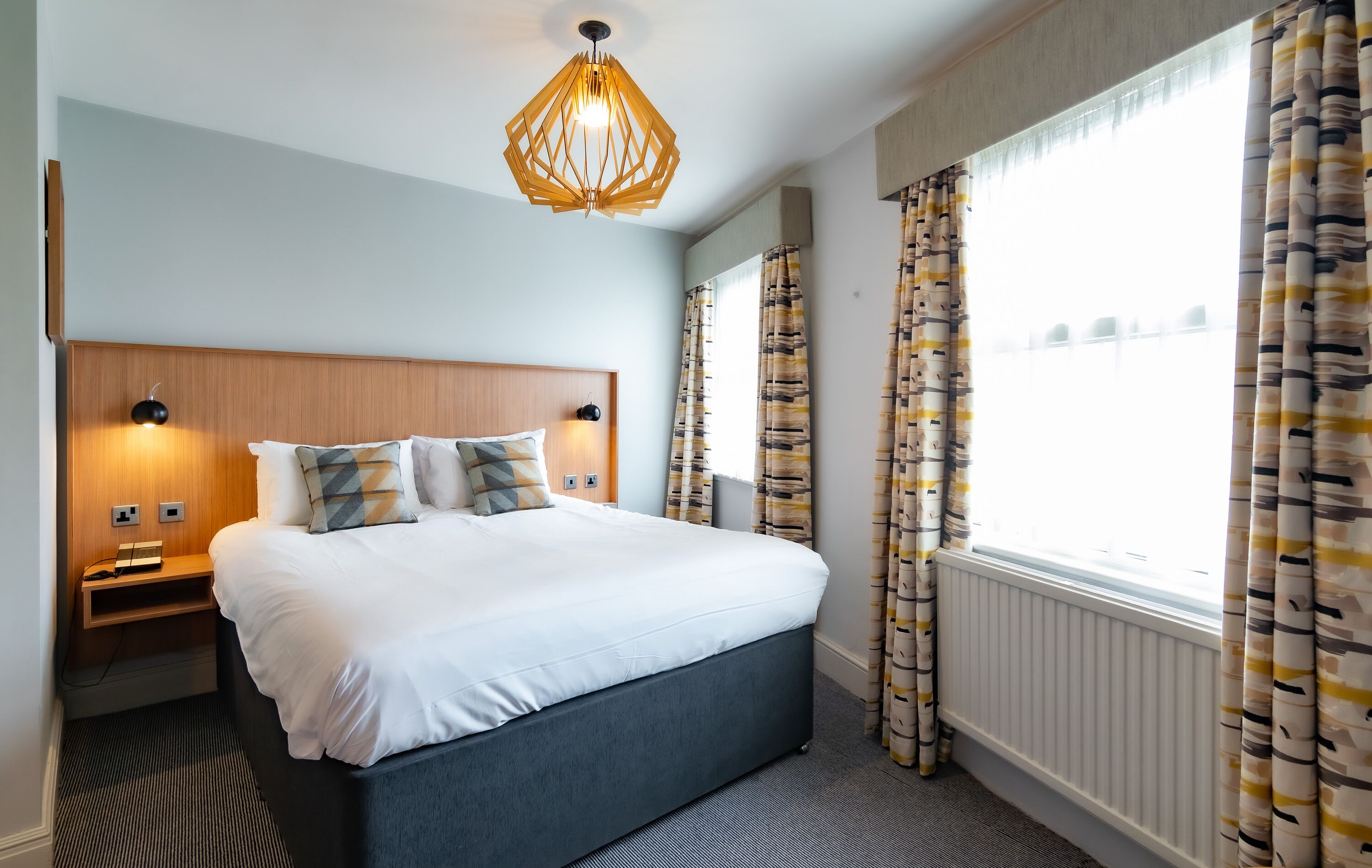 A modern hotel room with a large bed, a wooden headboard, bedside lamps, and a view of the city through large windows with patterned curtains.
