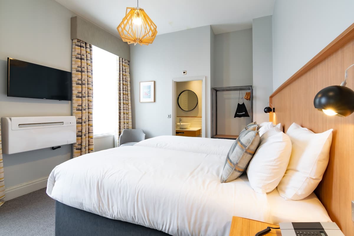 A modern hotel room with a large bed, a flat-screen TV mounted on the wall, a wooden headboard, and a bedside table with a lamp.