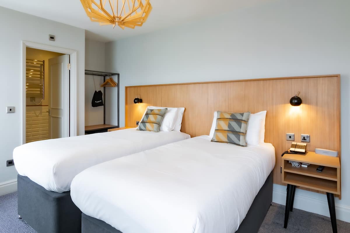 A hotel room with two beds, a wooden headboard, bedside lamps, a nightstand, and a coat rack.