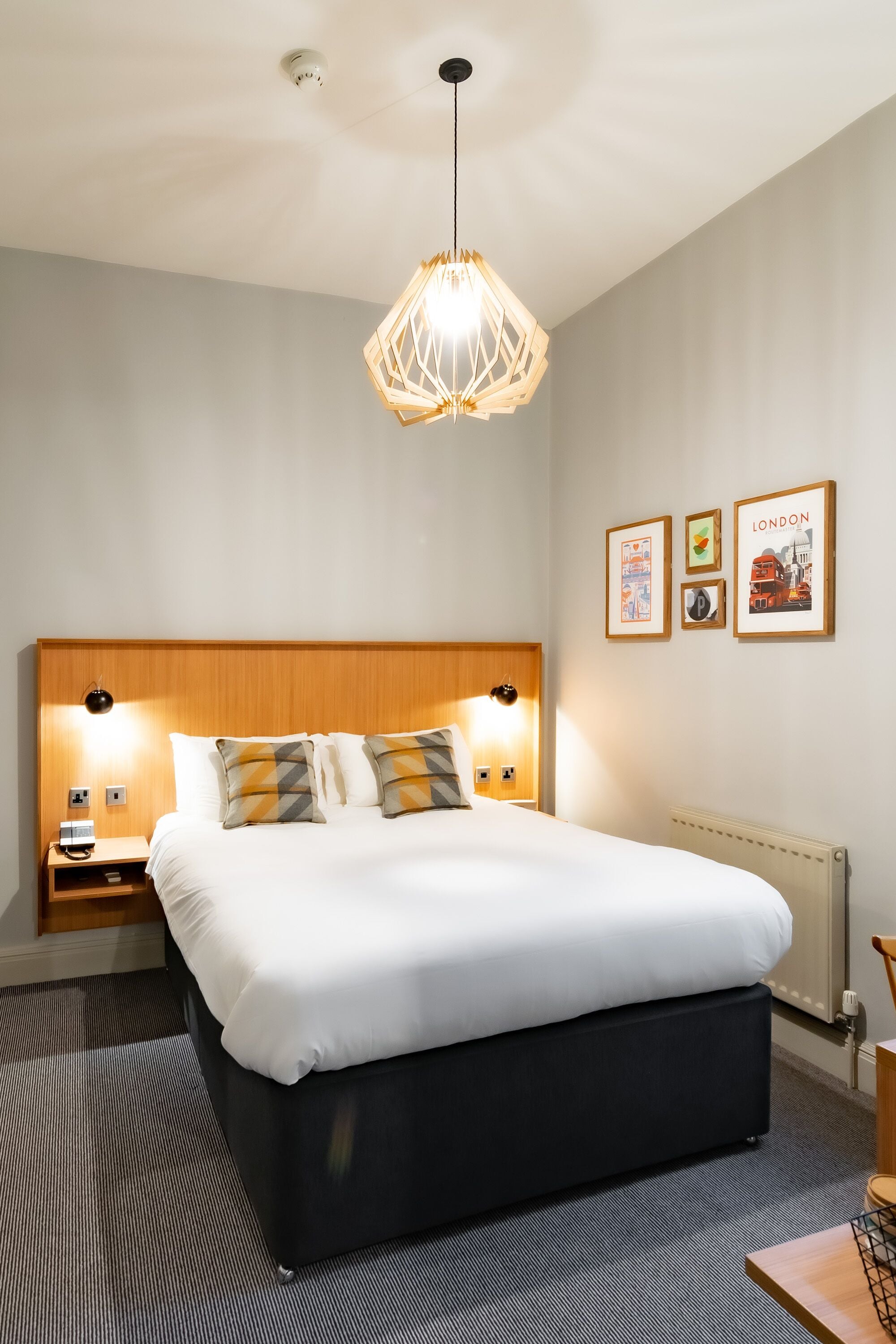 Una habitación de hotel moderna con una cama grande, cabecera de madera, una lámpara colgante geométrica singular y cuadros en la pared.