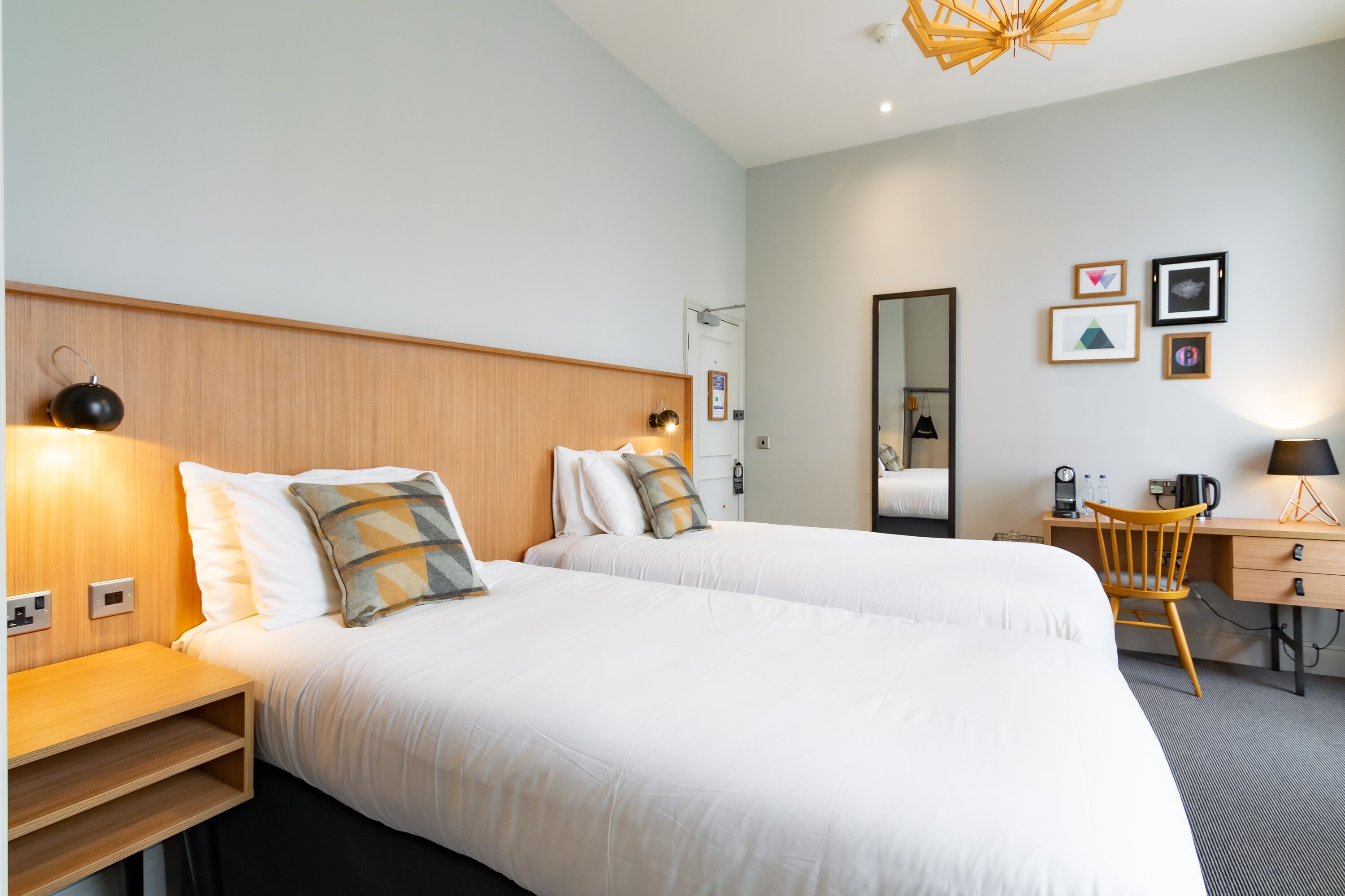 A hotel room with two beds, a desk, a chair, a mirror, and a wall with framed pictures.