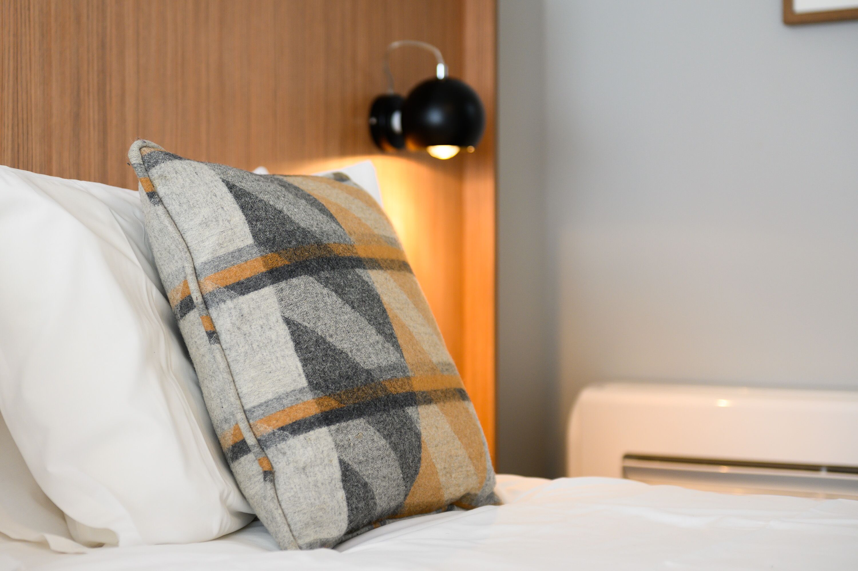 A bed with a plaid pillow and a plain pillow, a bedside table, and a wall-mounted lamp.