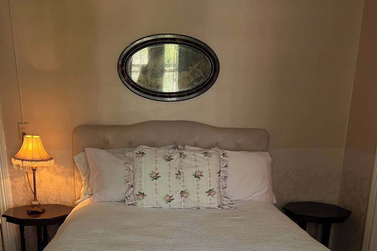 A neatly made bed with a white duvet, a ruffled skirt, and floral-patterned pillows. There is a round mirror on the wall above the bed and a table lamp on the left side.