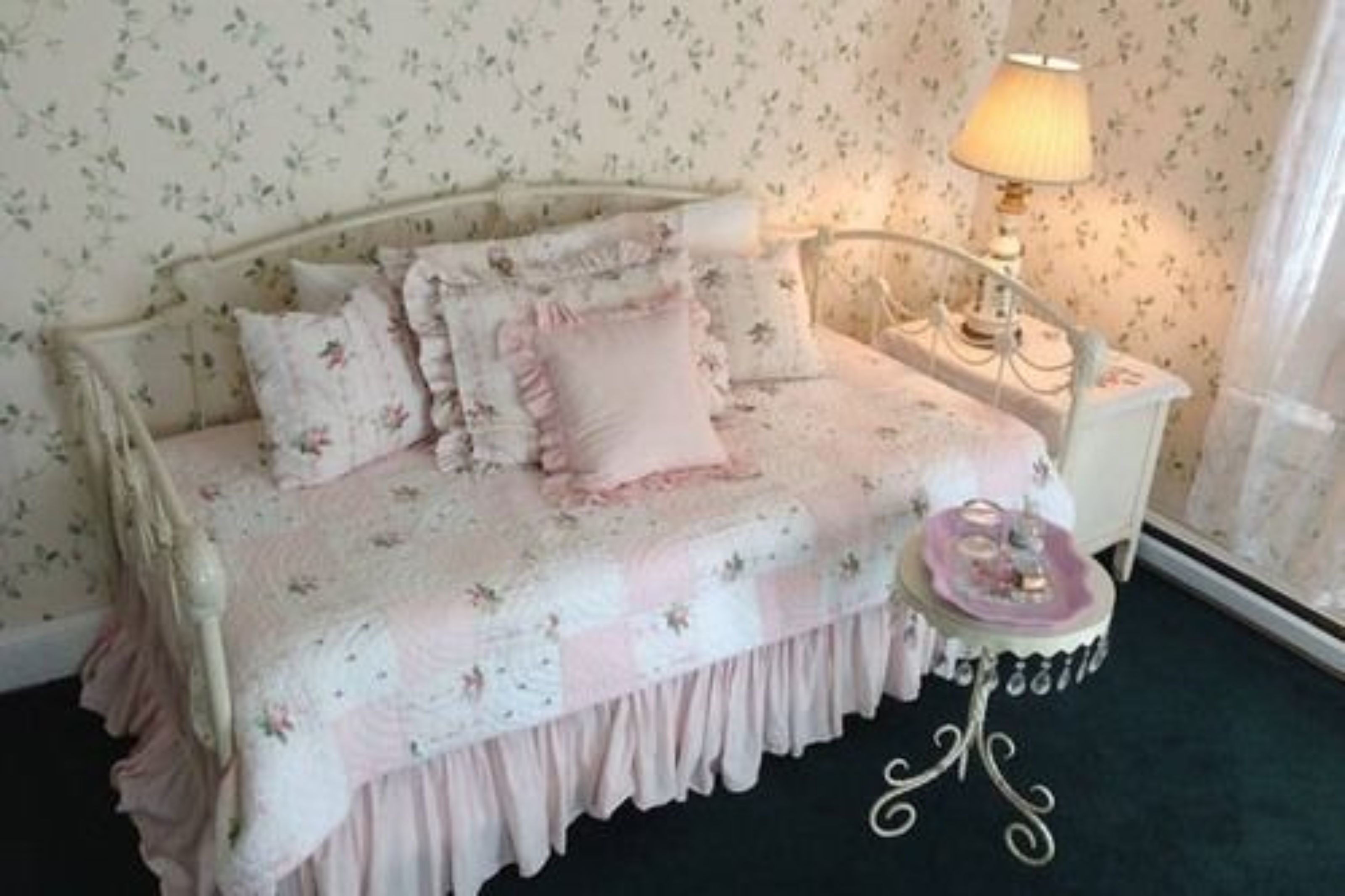 A bedroom with a floral-patterned bedspread, a bedside table with a lamp, and a window with curtains.